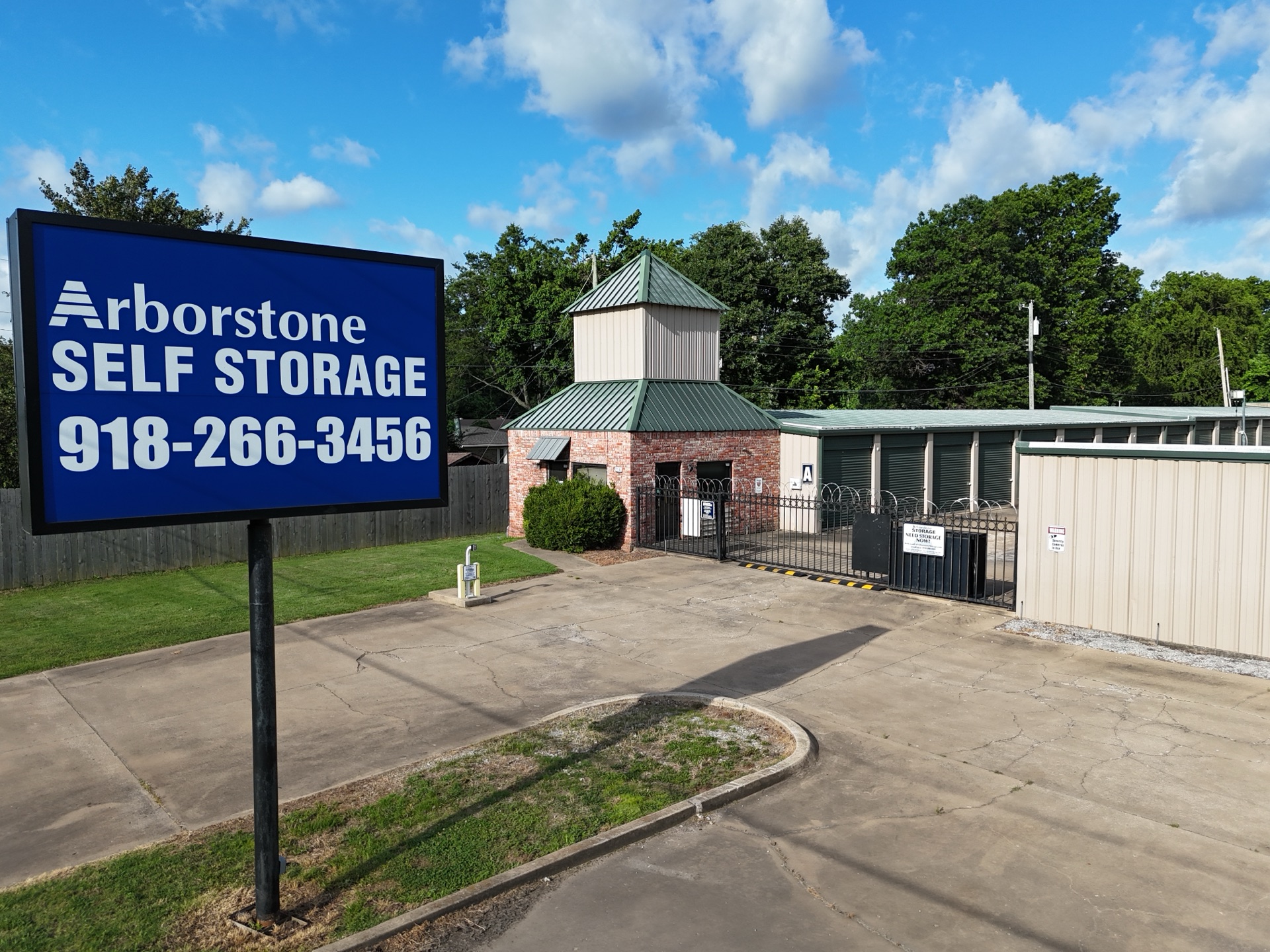 Arborstone Self Storage Catoosa entrance — large blue road sign with the Arborstone logo and 918-266-3456, next to a brick office building with a green metal cupola roof and secure black gate leading to drive-up storage units.