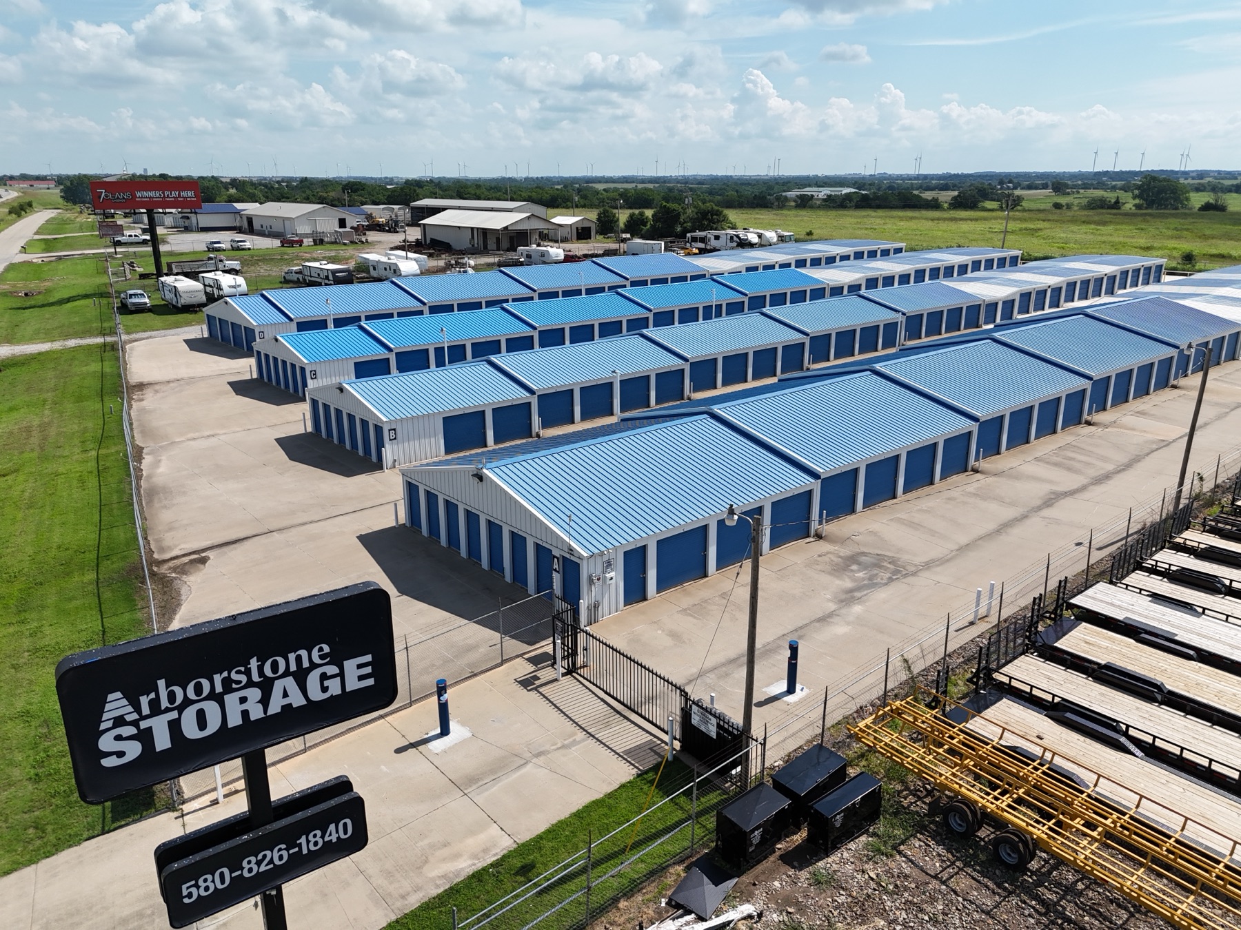 Aerial view of Arborstone Storage Ponca City, showing rows of drive-up units with blue roofs, wind turbines on the horizon, and the Arborstone Storage sign at the gate.