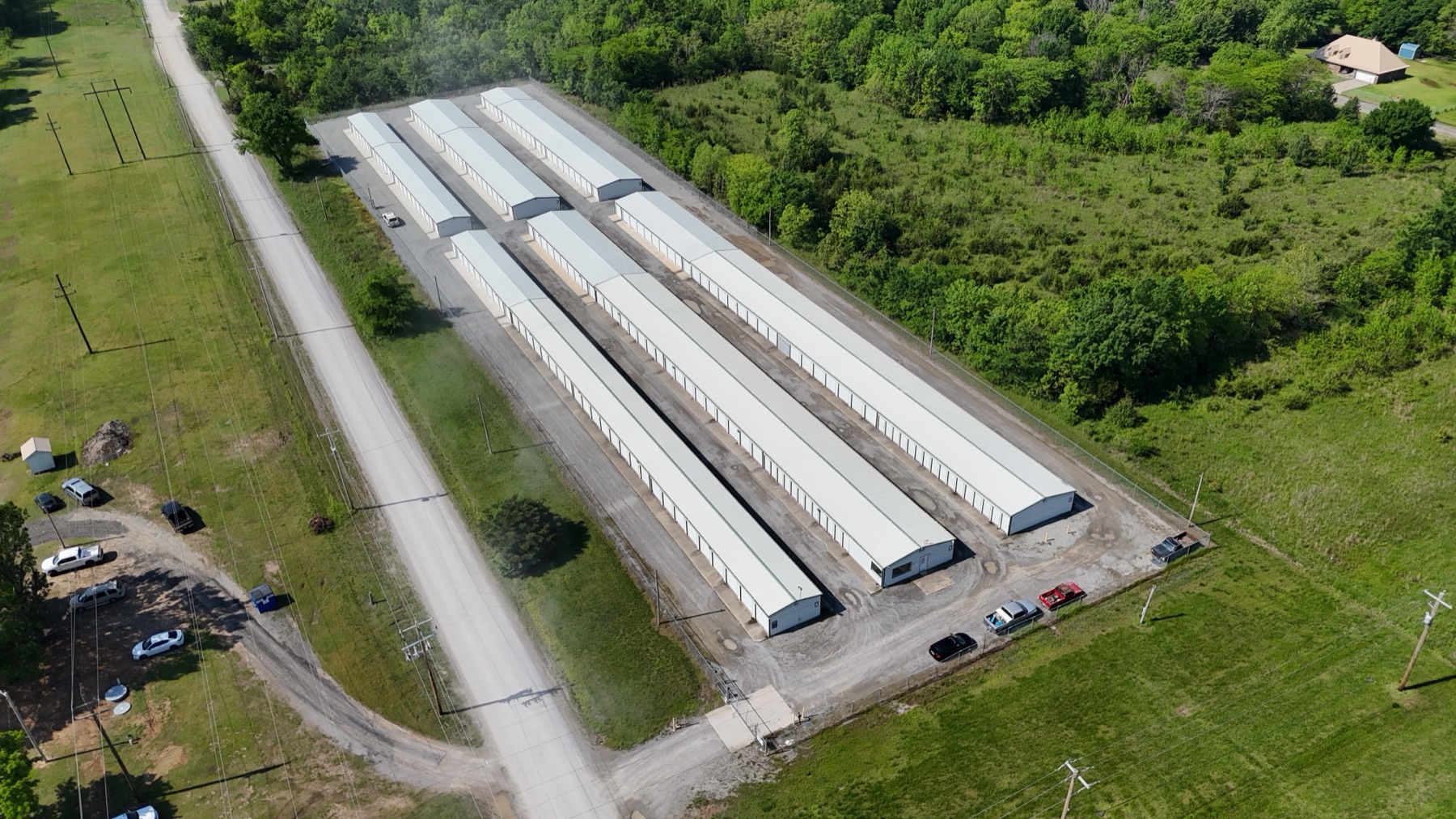Aerial view of Arborstone Storage Pryor, showing rows of drive-up units with white roofs surrounded by trees, with a dirt access road running alongside.