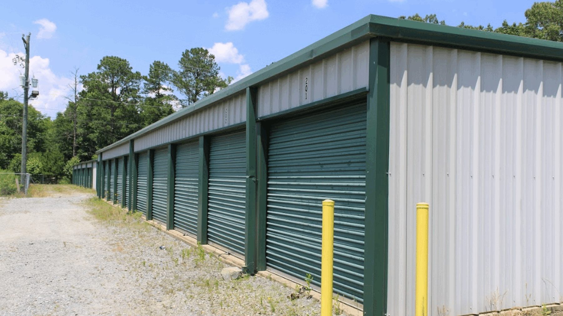 Arborstone Storage Mabelvale: a gravel drive aisle beside a row of drive-up storage units with green roll-up doors and yellow bollards.
