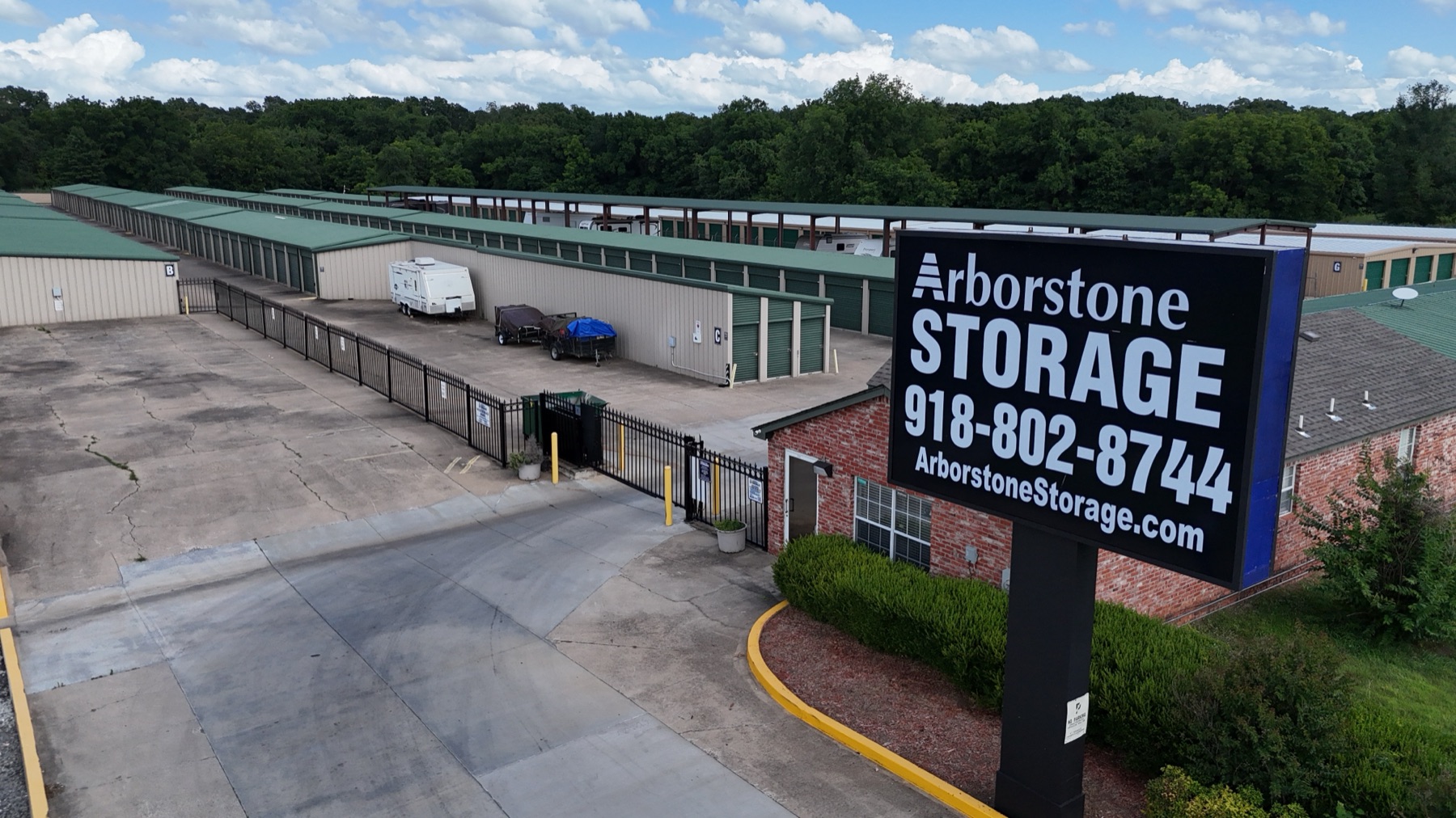 Aerial view of Arborstone Storage Tahlequah: the Arborstone Storage sign with phone number 918-802-8744 beside the office, the gated entrance and rows of green-roofed drive-up units with covered parking behind.