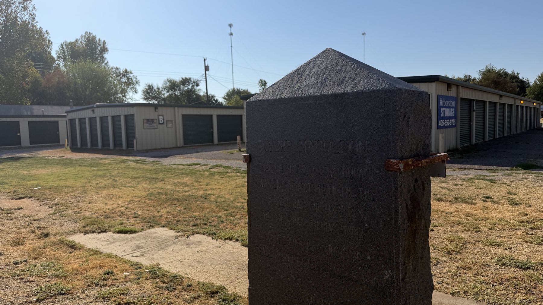 The granite Camp Napoleon marker on the grounds of Arborstone Storage Verden, with rows of drive-up storage units and an Arborstone Storage sign behind it.