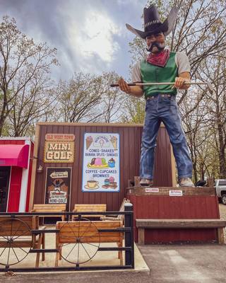 Jeb, a giant cowboy statue in a black hat, red neckerchief and green vest, holding a golf club beside the Old West Town Mini Golf sign in Hochatown.