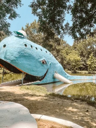 The smiling Blue Whale of Catoosa, a sky-blue concrete whale with porthole windows and a slide from its mouth, resting in a tree-shaded pond on Route 66.