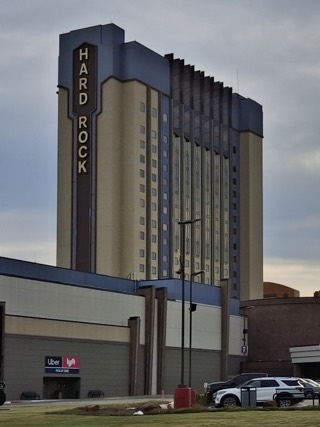 The Hard Rock Hotel & Casino Tulsa hotel tower, with a vertical Hard Rock sign, rising behind the casino building under a cloudy sky in Catoosa.