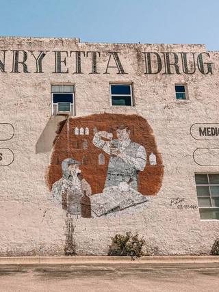 The whitewashed side wall of Henryetta Drug, with the store name painted in tall black letters above a hand-painted mural of a pharmacist pouring medicine beside a customer.