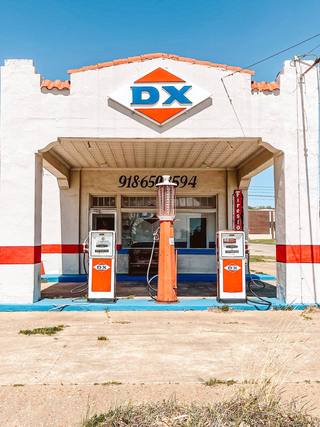 A restored white filling station with a red tile roofline, a blue-and-orange DX diamond sign, and vintage orange DX gas pumps under its canopy in Henryetta.