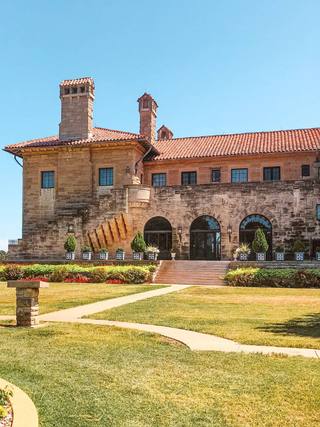 The sandstone Marland Mansion with its red tile roof, tall chimneys and arched doorways, seen across a green lawn and flower beds under a clear blue sky.