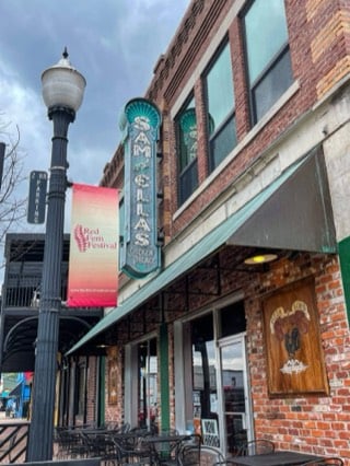 Sam & Ella’s Chicken Palace on N. Muskogee Avenue in downtown Tahlequah: a red-brick storefront with a vintage vertical sign and green awning, beside a lamppost with a Red Fern Festival banner.