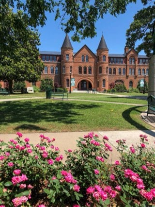 Seminary Hall at Northeastern State University in Tahlequah: a red-brick Romanesque Revival building with twin conical towers and an arched entry, seen across the lawn past a bed of pink roses.